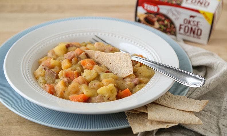 Enkel lapskaus med bacon og grønnsaker servert i hvit skål på lyseblå dekketallerken. Skålen står på lyst trebord med linserviett og flatbrød på siden, og en eske Toro oksefond i bakgrunnen.