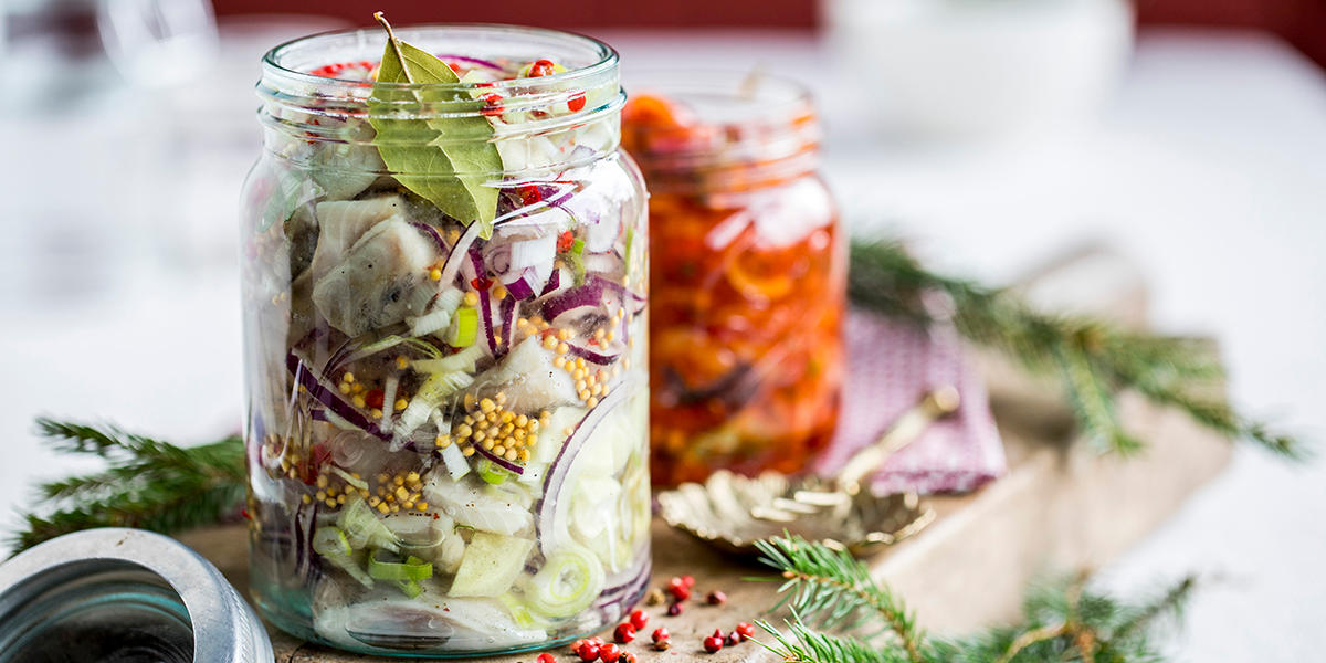 Hjemmelaget sursild på Norgesglass med sennepfrø, rødløk i skiver, krydder og urter. Norgesglasset med sursild står ved siden av et mindre Norgesglass med hjemmelaget tomatsild på rustikk trefjøl med granbar og tørkede krydder.