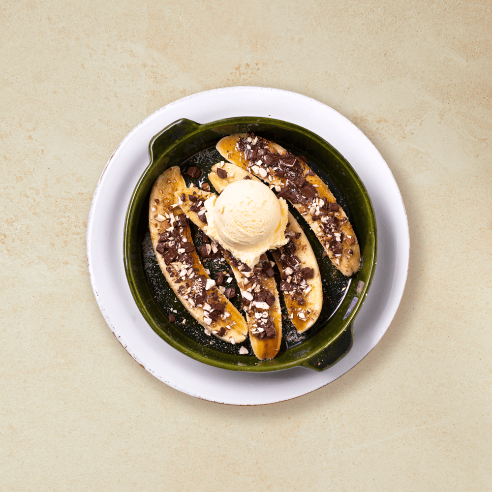 Ovnsbakt banan skivet på langs, med brunt sukker, sjokolade og hakkede mandler, servert i rund grønn ildfast form med en kule vaniljeis på toppen.