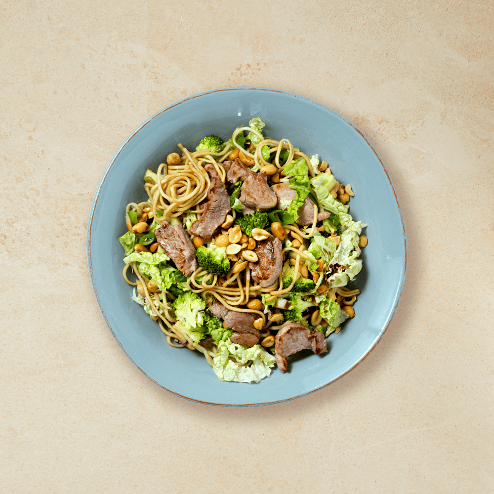 Asiatisk nudelsalat med svinekjøtt, servert med peanøtter i lyseblå skål.