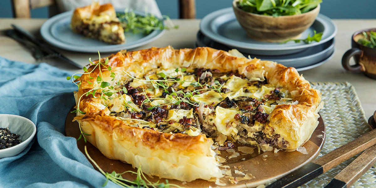 Pai med sprø bunn fylt med sopp, bacon og ost og toppet med frisk timian. Det er skåret et stykke av paien. 