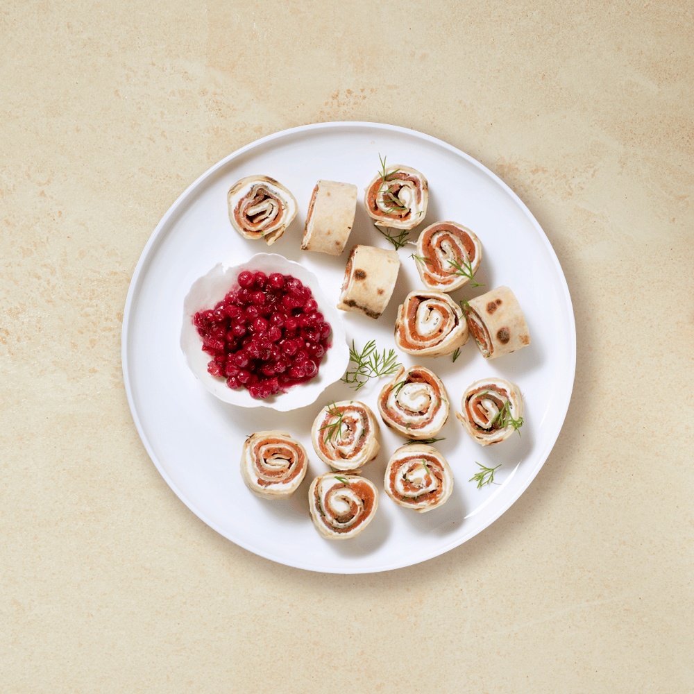 Lefserull med røkelaks, kremost og dill, skåret i skiver og servert på hvit tallerken med en hvit skål tyttebær ved siden av. Det er drysset fersk dill over lefserullene.