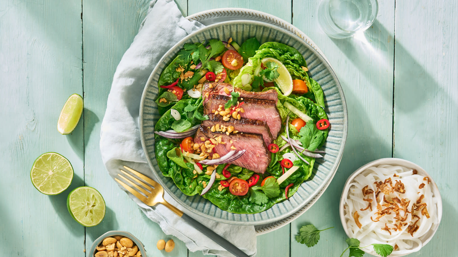 Klassisk thai biffsalat med rosastekt kjøtt, rødløk, tomater, agurk, lime og salte peanøtter servert i blå skål. Liten skål med glassnudler, og liten skål med peanøtter på siden, og frisk lime rundt.