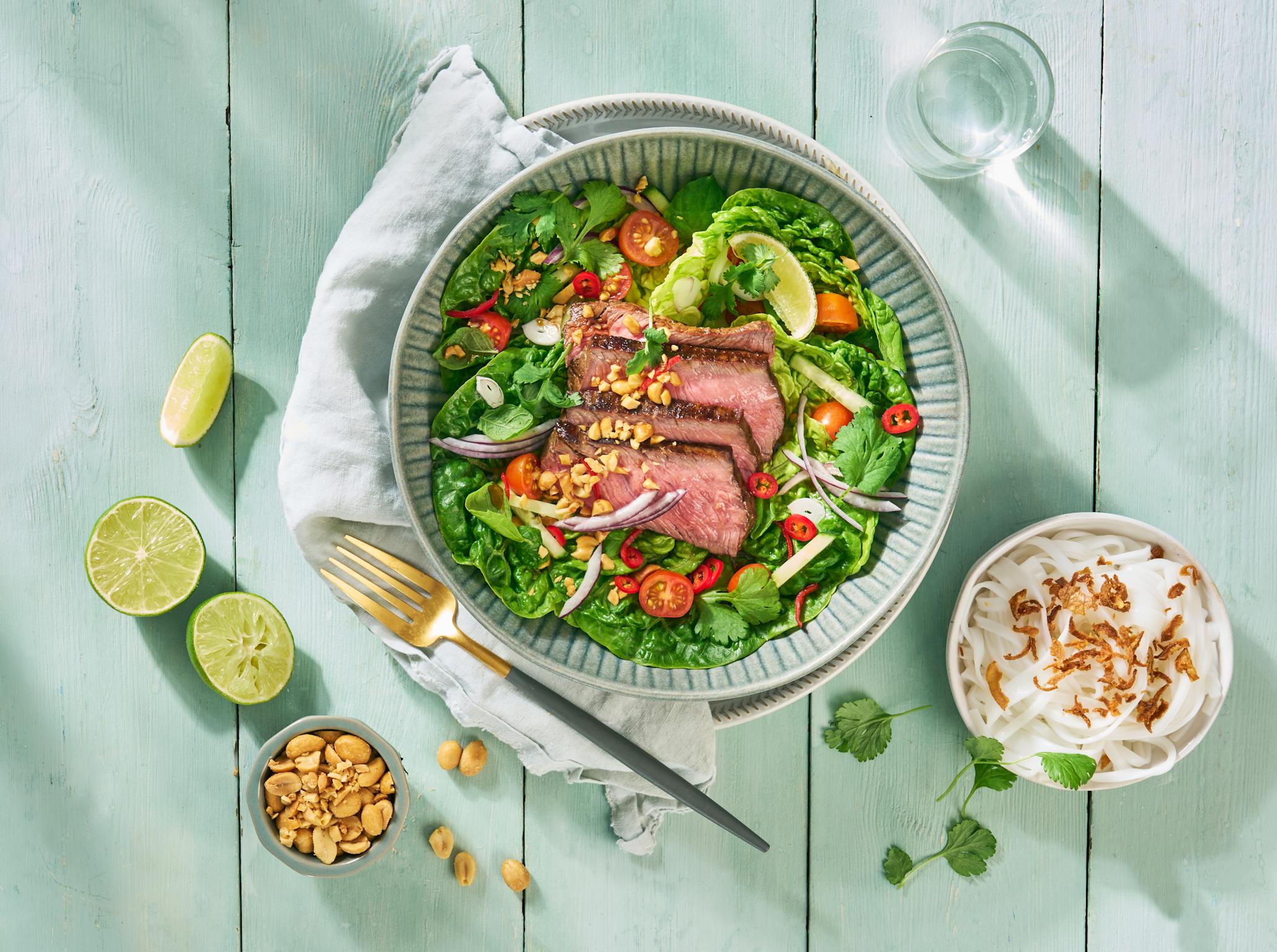 Klassisk thai biffsalat med rosastekt kjøtt, rødløk, tomater, agurk, lime og salte peanøtter servert i blå skål. Liten skål med glassnudler, og liten skål med peanøtter på siden, og frisk lime rundt.