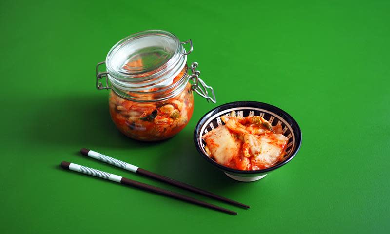 Hjemmelaget kimchi, fermentert kål og andre grønnsaker, på syltetøyglass. Ved siden av står en skål med kimchi, ved siden av et par spisepinner. Grønn bakgrunn.