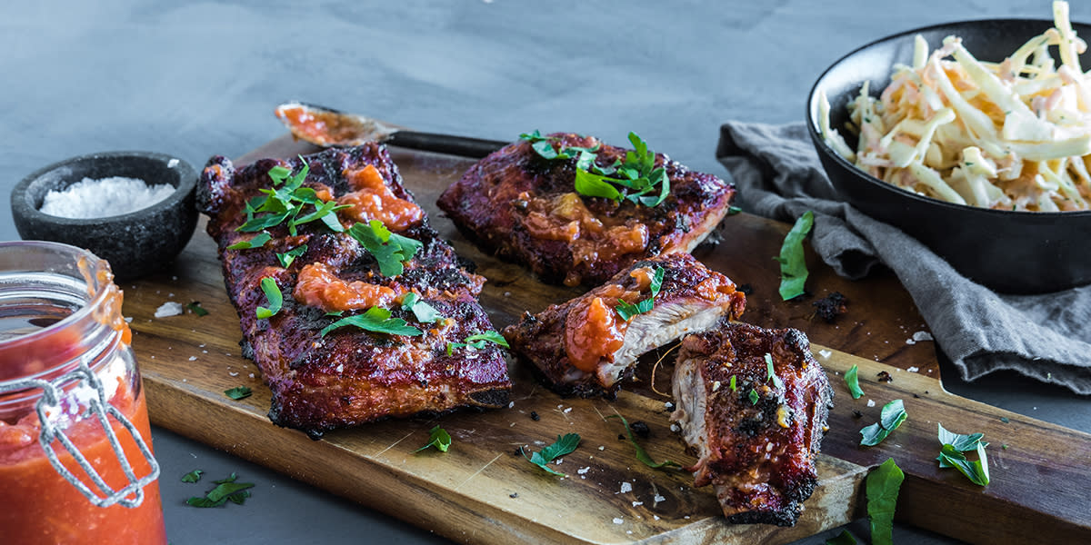 Spareribs med hjemmelaget rub og mango bbq-saus servert på trefjøl med saus og friske urter på toppen. Svart skål med coleslaw på mørk stoffserviett ved siden av.