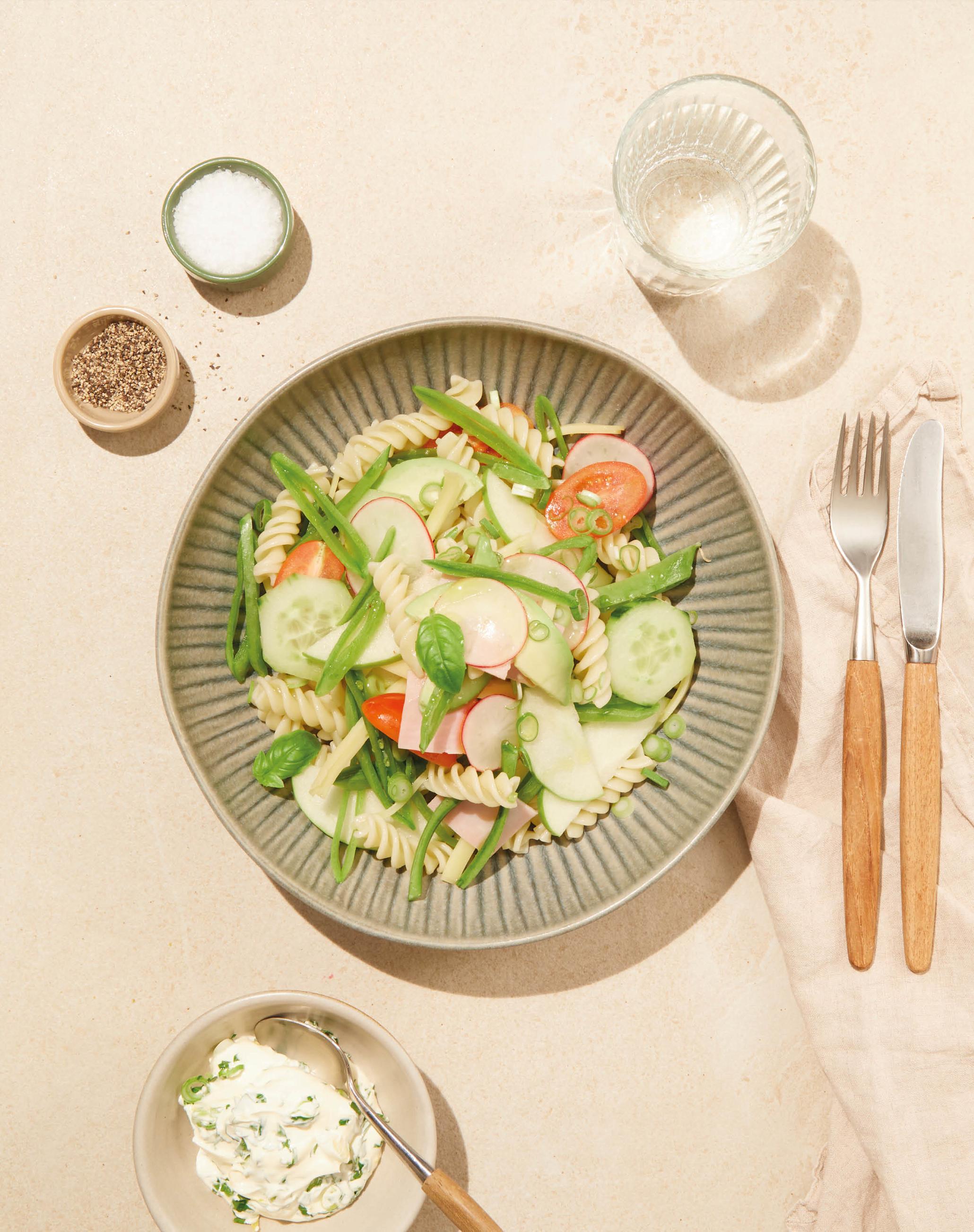 Fargerik pastasalat med ost, skinke, tomat, agurk, avokado og reddik i lys bolle på et bord med lys duk.