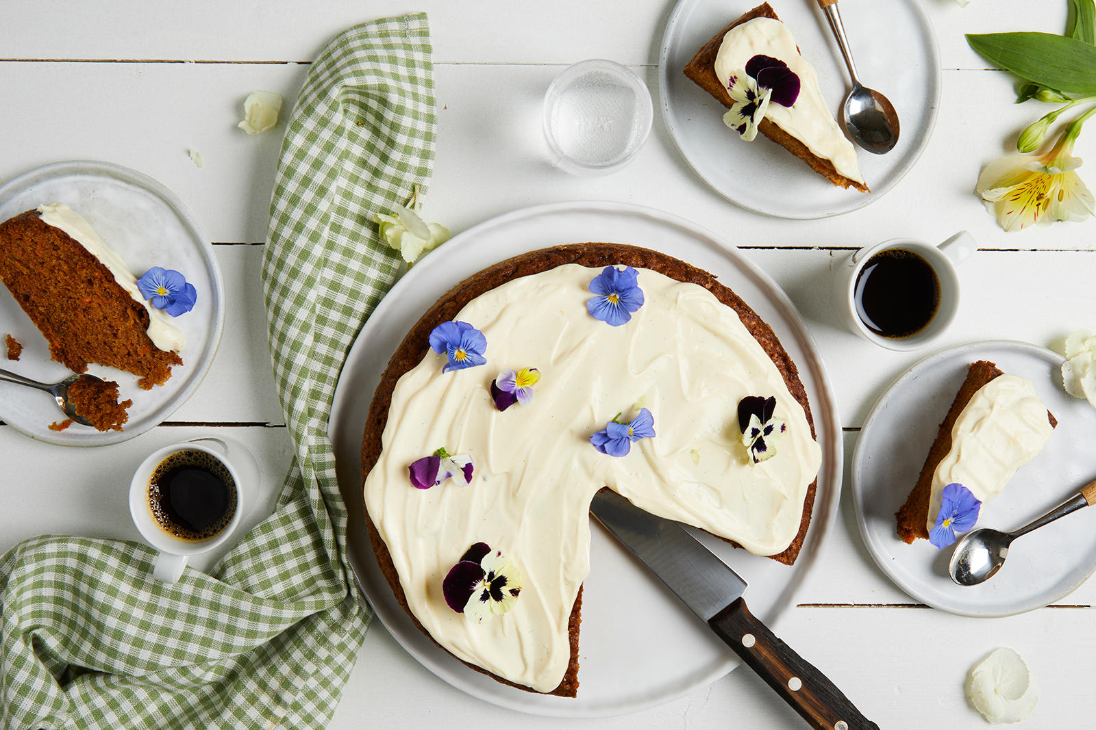 Krydret gulrotkake med ostekrem pyntet med stemorsblomster i gult og lilla, og servert på lyst rundt kakefat på hvitt trebord, med kaffekopper og asjetter med kakestykker.