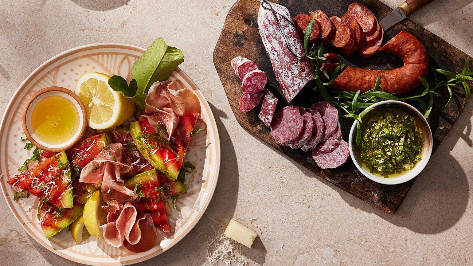 Grillet vannmelon med serrano og manchego servert på mønstret lyst fat med olivenolje, sitronbåter og friske urter. Fat med spekemat ved siden av.