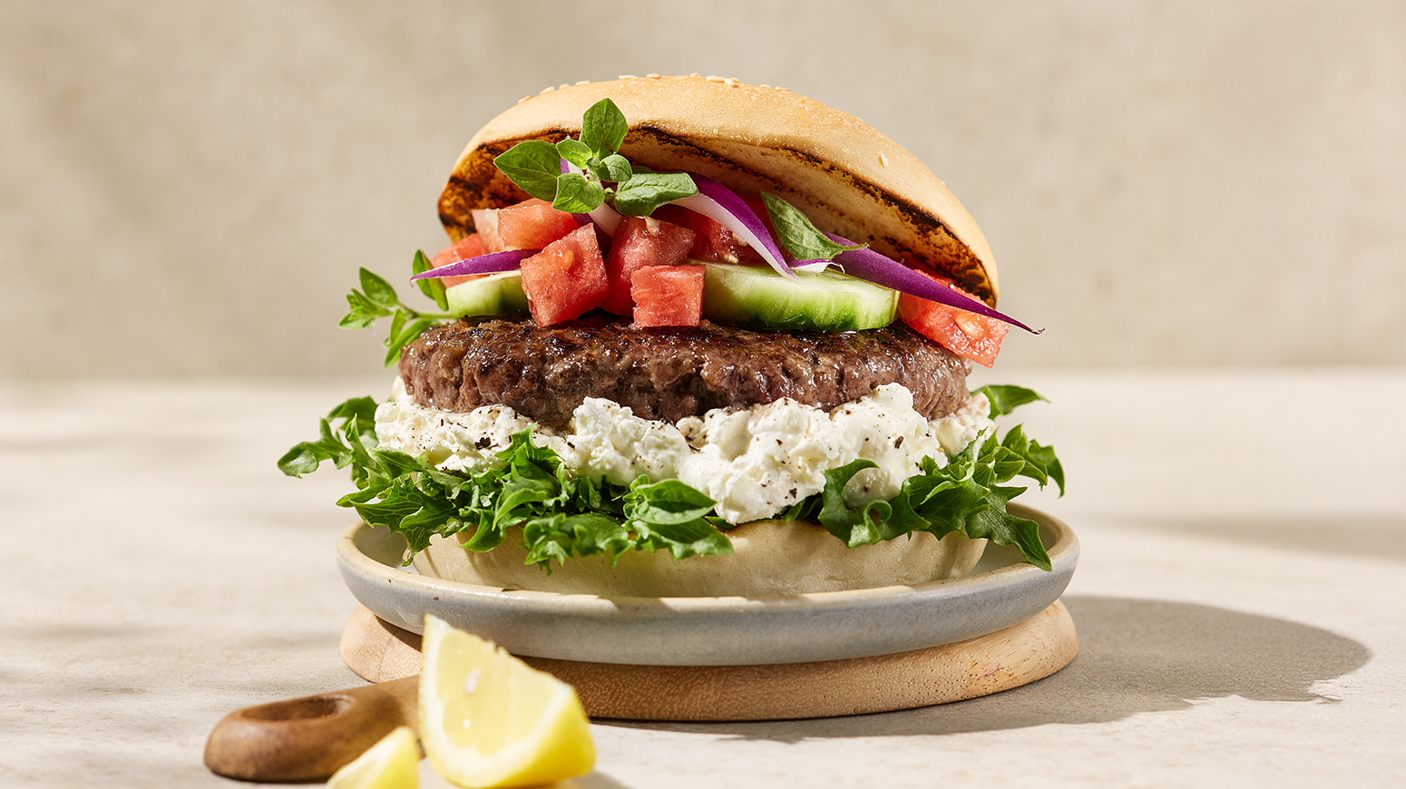 Gresk burger med vannmelonsalat og fetaostkrem i burgerbrød servert på lys tallerken på lys trefjøl, med sitronbåter i forgrunn.