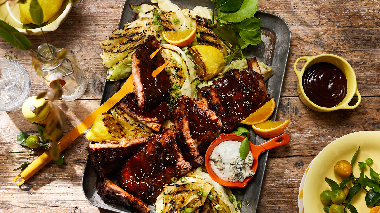 Spareribs med grillet nykål og Blue Cheese-dressing servert på metallfat med halve grillede sitroner. Skåler med tilbehør rundt.