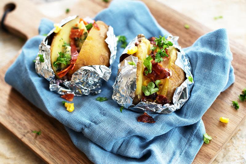 To bakte poteter i aluminiumsfolie ligger på et blått linhåndkle, på rustikk trefjøl. Potetene er fylt med hjemmelaget kryddersmør, bacon, mais, og finhakket persille.