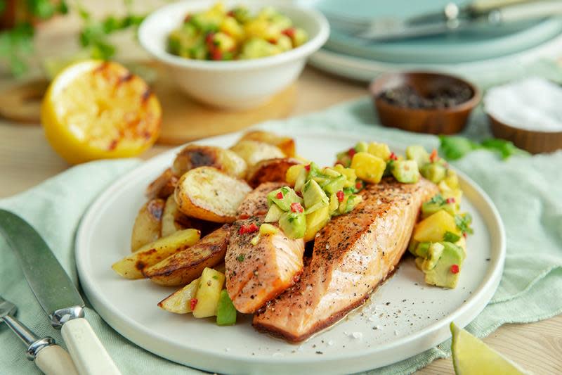 Grillet laks med eksotisk avokado- og mangosalsa servert på lys flat tallerken med ovnsbakte poteter. Liten skål med avokado- og mangosalsa i bakgrunnen, med en halv grillet sitron ved siden av.