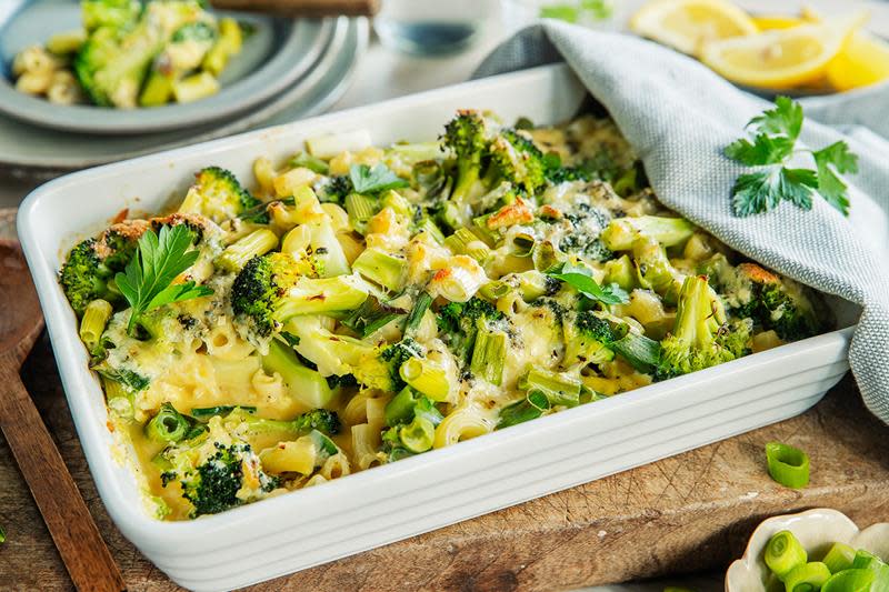 Frisk og grønn brokkoligrateng med makaroni og ost, servert i hvit ildfast form, på trefjøl. Lyst blått linhåndkle dandert over, med sitronbåter på siden.