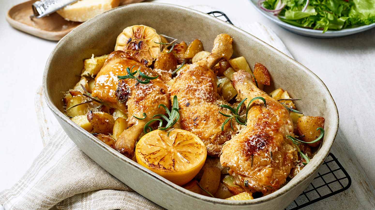 En grå, oval ildfast form med fire stykker ovnsbakt kyllinglår og potetbiter og hvitløksfedd og en halv, grillet sitron. Formen står på en rist på et hvitt kjøkkenhåndkle. I bakgrunnen er en skål med grønn salat og et fat med en bit parmesanost.