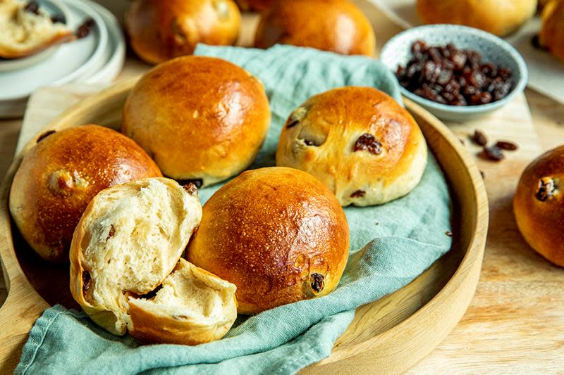 Gylne hveteboller med rosiner bakt med Tangzhong metoden, servert på trebrett med grønt linhåndkle. Liten skål med rosiner ved siden av, omkranset av nybakte boller.