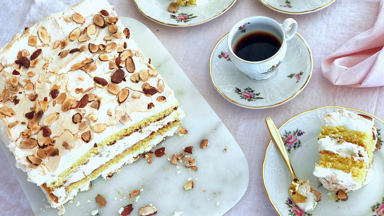 Klassisk Kvæfjordkake servert på hvitt marmorfat, på lys duk. Småblomstret kaffeservise med kaffekopper, og asjetter med biter av kake.