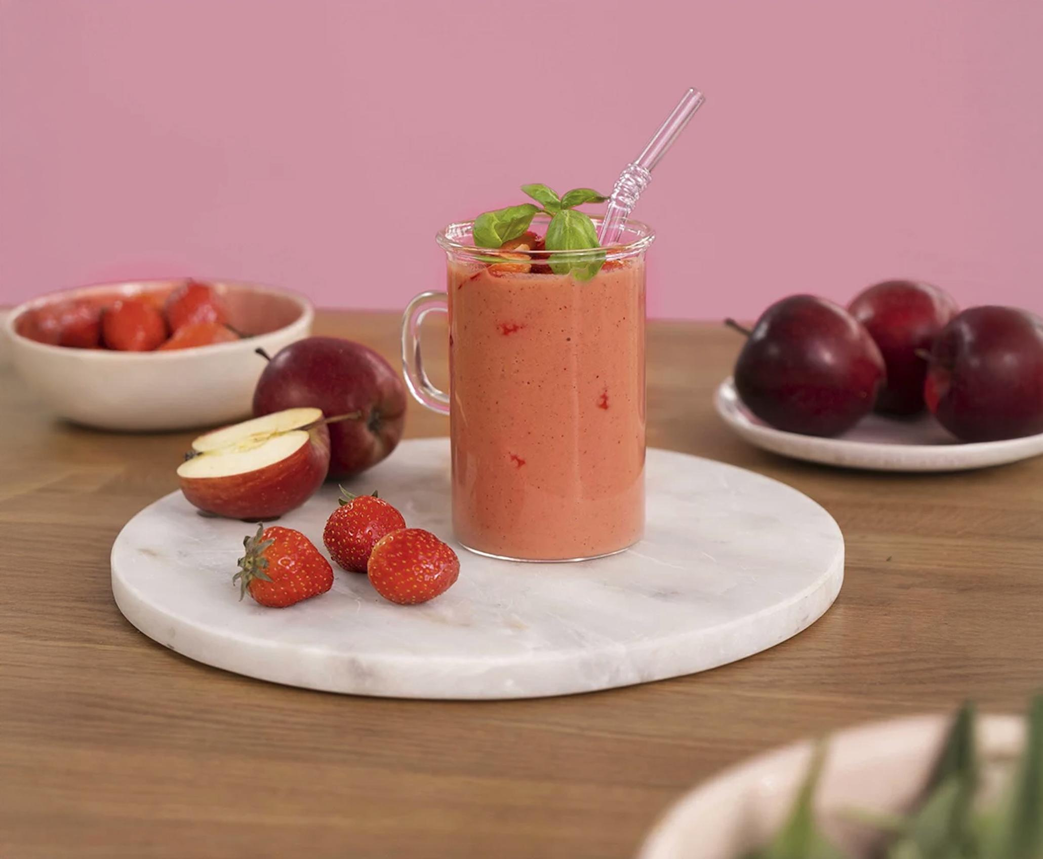 Rosa smoothie med eple, jordbær og basilikum i høyt gjennomsiktig krus, på hvitt marmorfat på trebord. Ferske jordbær og epler i bakgrunnen.