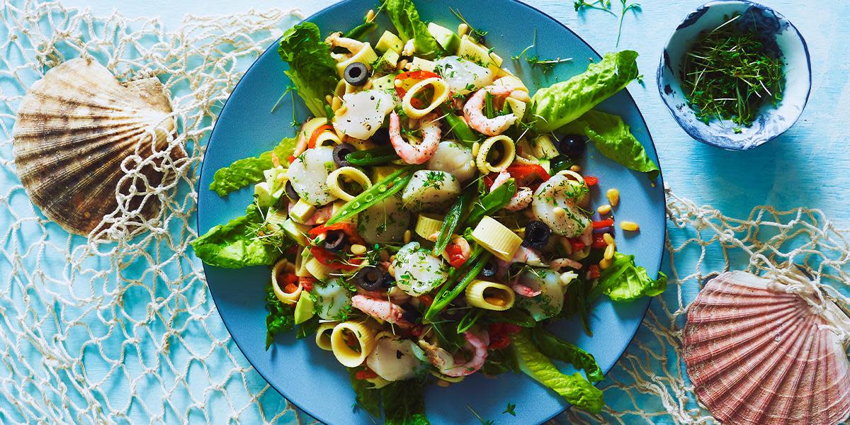Pastasalat med kamskjell og håndpillede reker, grønnsaker og salat på blå tallerken.