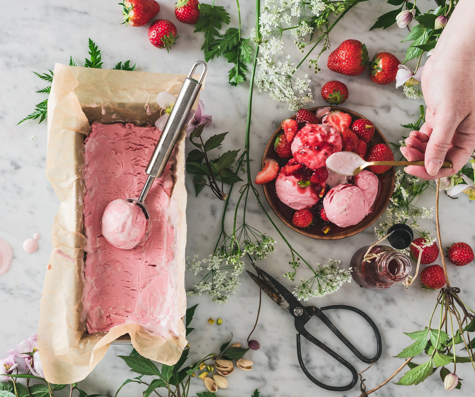 Hjemmelaget jordbæris i bakepapirkledd brødform, på lys marmorbakgrunn. Grønne planter, markblomster og ferske bær rundt. Skål med flere kuler jordbæriskrem og ferske jordbær på siden. En hånd holder en skje over skålen.