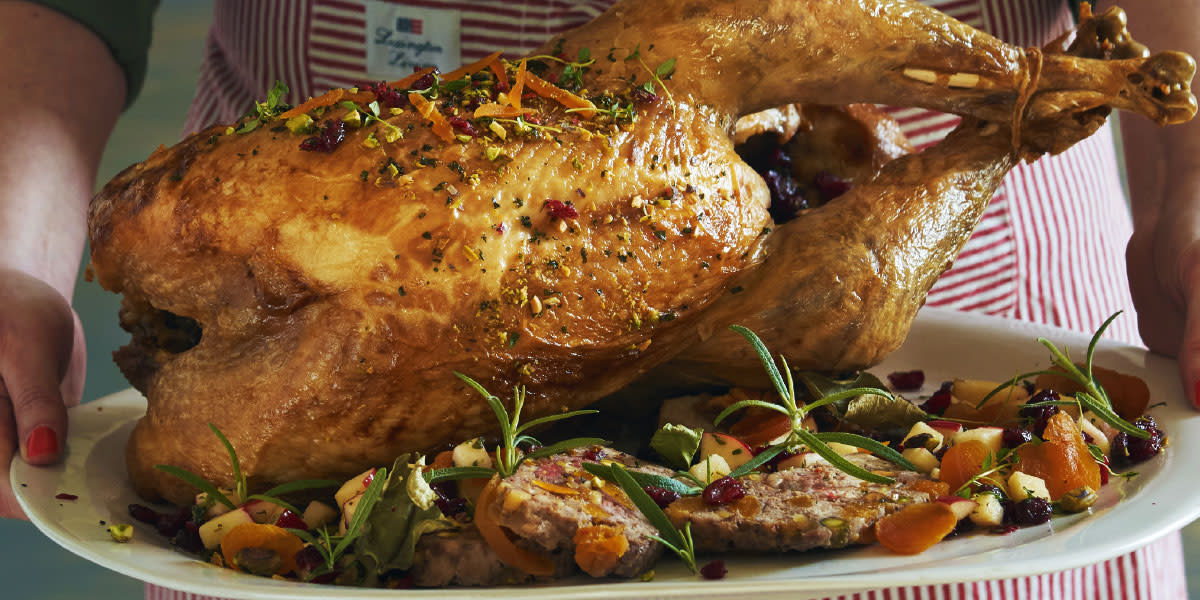 Helstekt kalkun med kjøttstuffing og hjemmelaget fløtesaus servert på hvitt ovalt fat, med skiver av stuffing, tørket frukt og fersk rosmarin. Fatet holdes av to kvinnehender.