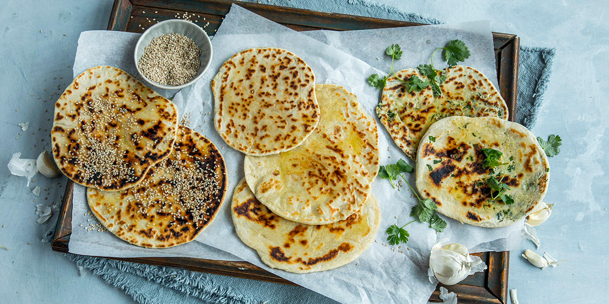 Nanbrød på tre måter servert på trefat med bakepapir. Hel hvitløk, friske urter og liten skål med sesamfrø ved siden av.