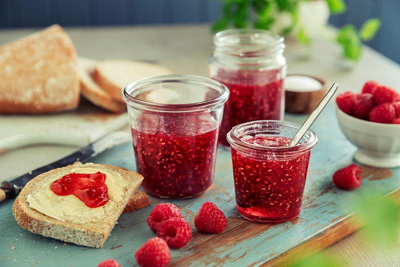 Knallrødt bringebærsyltetøy på forskjellige glass, står på blåmalt treplanke. En brødskive med smør og rødt syltetøy står ved siden av.