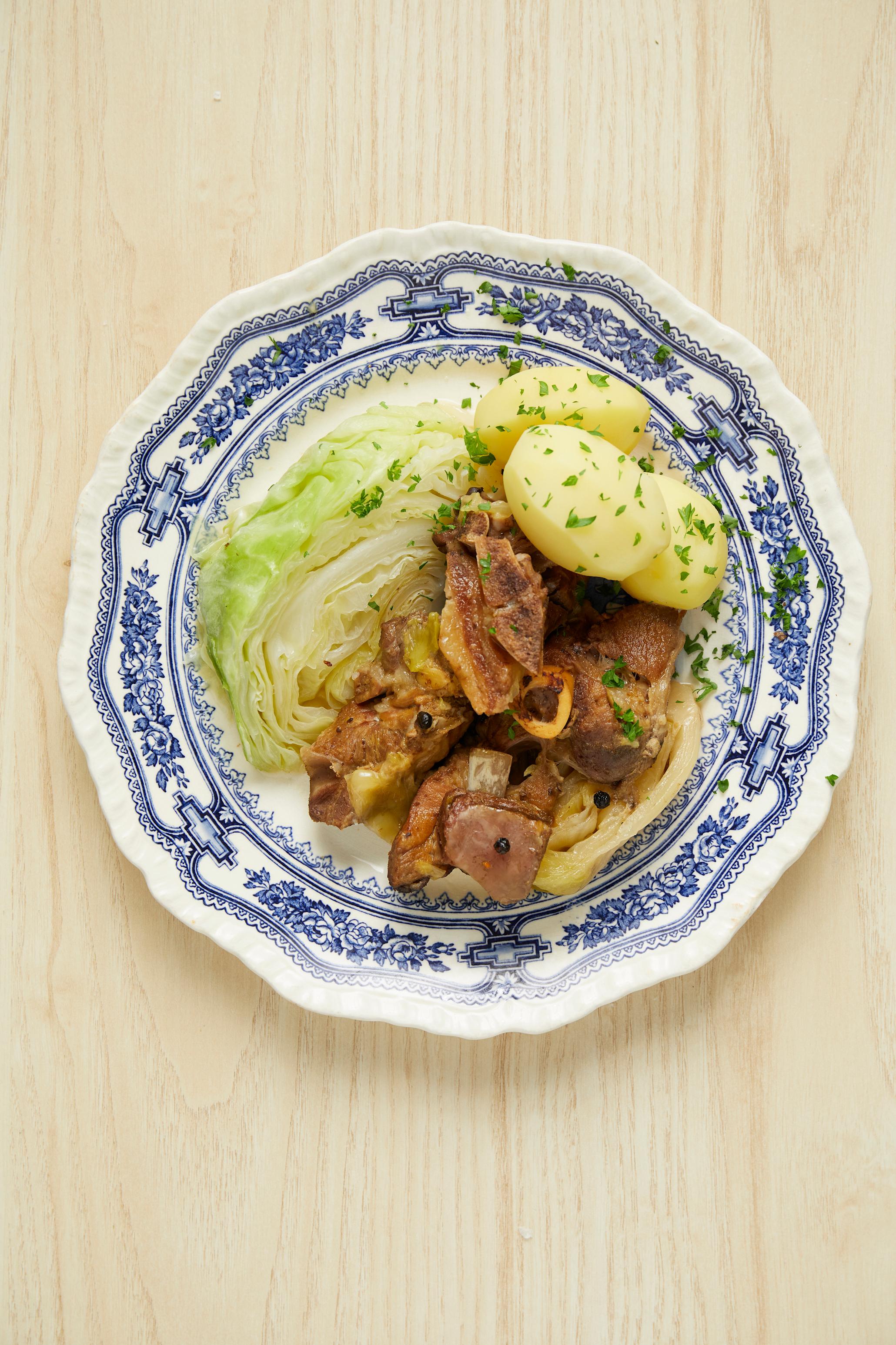 Fårikål med hvitløk, syrlig eplejuice og toppkål, servert med kokte poteter og et dryss persille på blåmønstrert hvit tallerken, på lyst trebord.