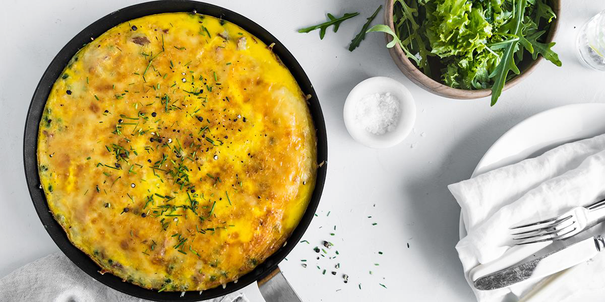 Bondeomelett med potet og skinke, pyntet med gressløk og servert i svart støpejernspanne på hvitt bord. Skål med blandet salat og dekketøy ved siden av.