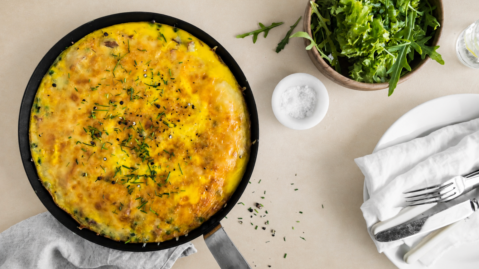 Bondeomelett med potet og skinke, pyntet med gressløk og servert i svart støpejernspanne på beige bakgrunn. Skål med blandet salat og dekketøy ved siden av.