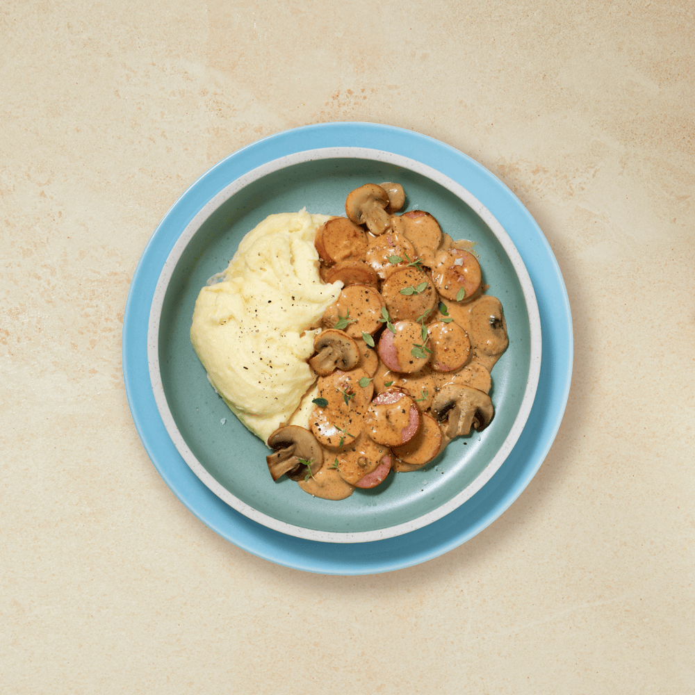 Stroganoff med pølse, sopp og potetmos, servert på en turkis tallerken på beige bakgrunn.