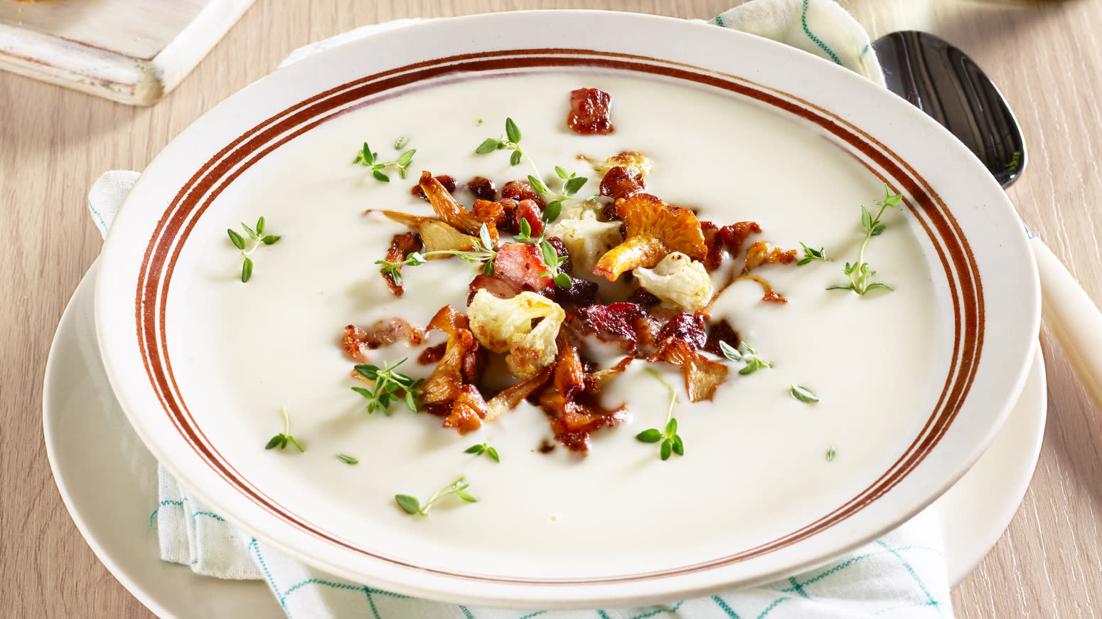 Blomkålsuppe med bacon og kantareller servert med et dryss frisk timian i hvit skål med røde striper rundt kanten. Spiseskje på siden.