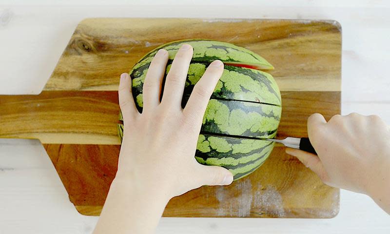 En halv vannmelon ligger på trefjøl mens to hender deler den opp i biter med en kniv.