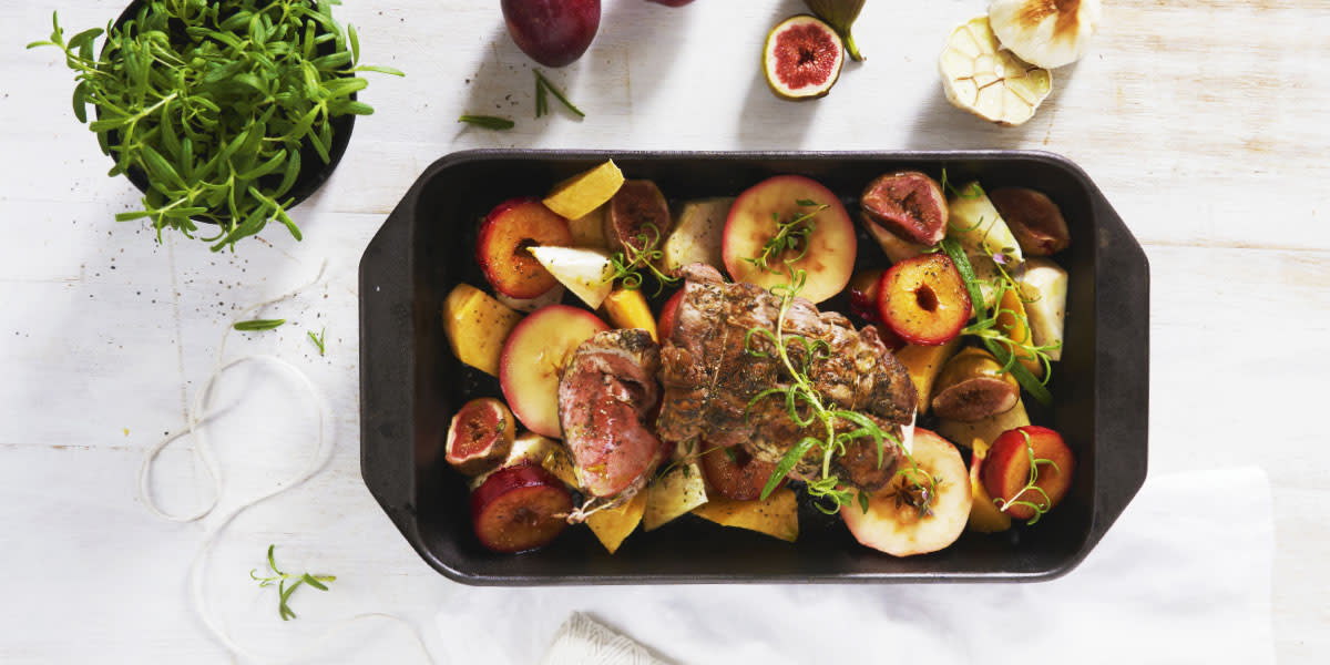 Lammestek med ovnsbakte frukter, servert i svart ildfast form med ferske urter over. Formen står på hvitt trebord.