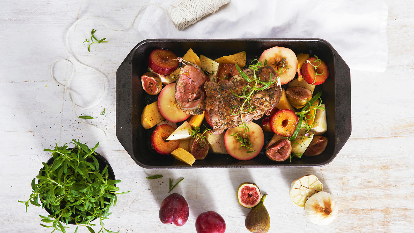 Lammestek med ovnsbakte frukter, servert i svart ildfast form med ferske urter over. Formen står på hvitt trebord.