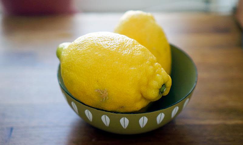 To sitroner ligger i en grønn bolle med hvitt mønster, på et trebord.