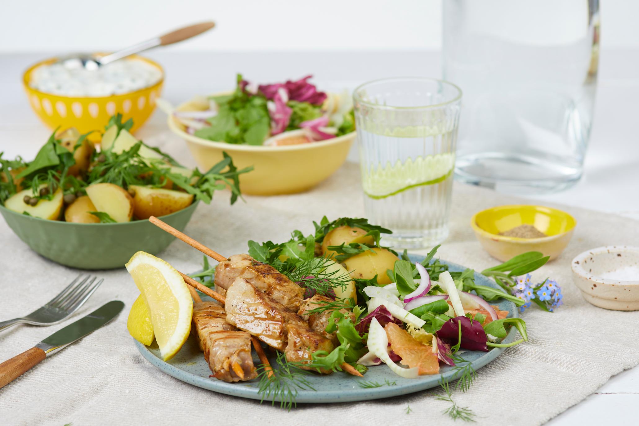 Grillspyd Spicy Lemon med grapefruktsalat på en turkis tallerken foran en grønn skål med poteter og 2 gule skåler med dressing og salat