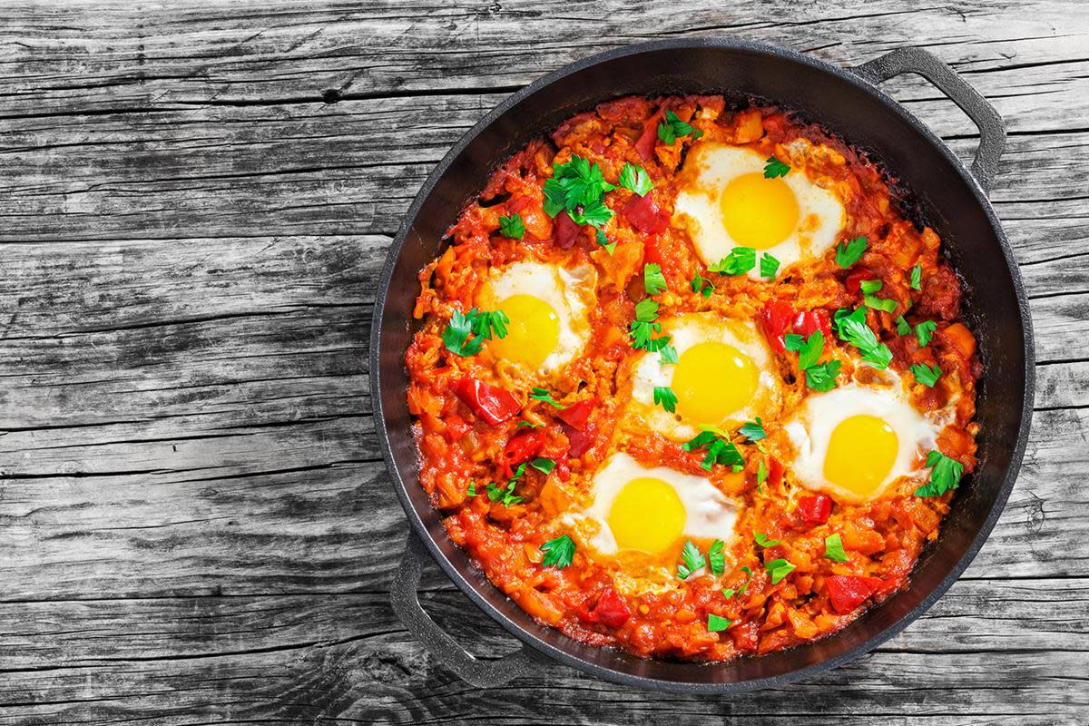 Shakshouka med bakte egg i tomatsaus, servert med urtedryss i støpejernsgryte på rustikk trebord.