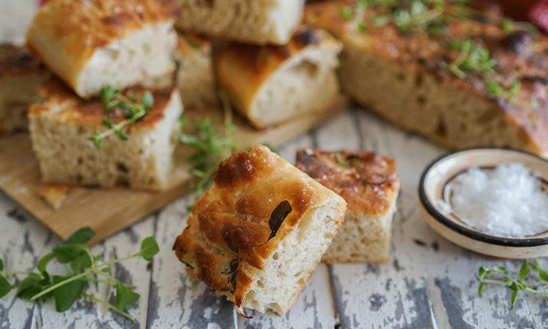 Enestående Mats eltefrie focaccia, ferdig stekt og delt opp i biter. Servert på rustikk hvitt trebord, med ferske urter og en liten skål med salt.