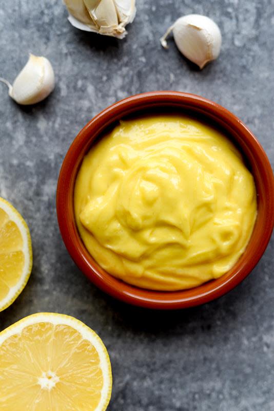 Hjemmelaget aioli i keramikkbolle på grå bakgrunn. Sitronskiver og hvitløksfedd på siden.