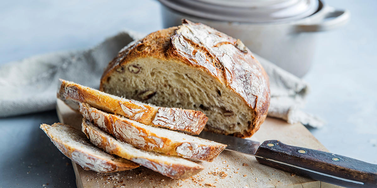 Saftig eltefritt grytebrød med pekannøtter skåret i skiver og servert på trefjøl med rustikk brødkniv. Lys kjele og linserviett i bakgrunnen.