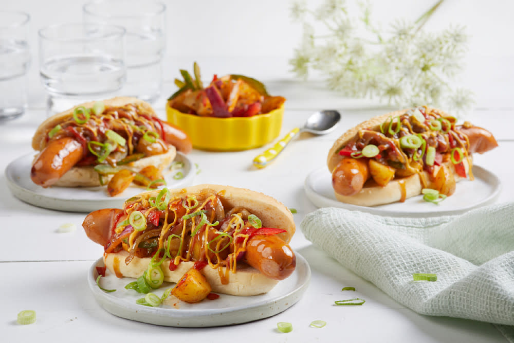 Cheesy Bacon pølse med karamellisert løk og potetsalat servert i pølsebrød på hvite små asjetter, på hvitt trebord. Gul skål med fargerik potetsalat i bakgrunnen.