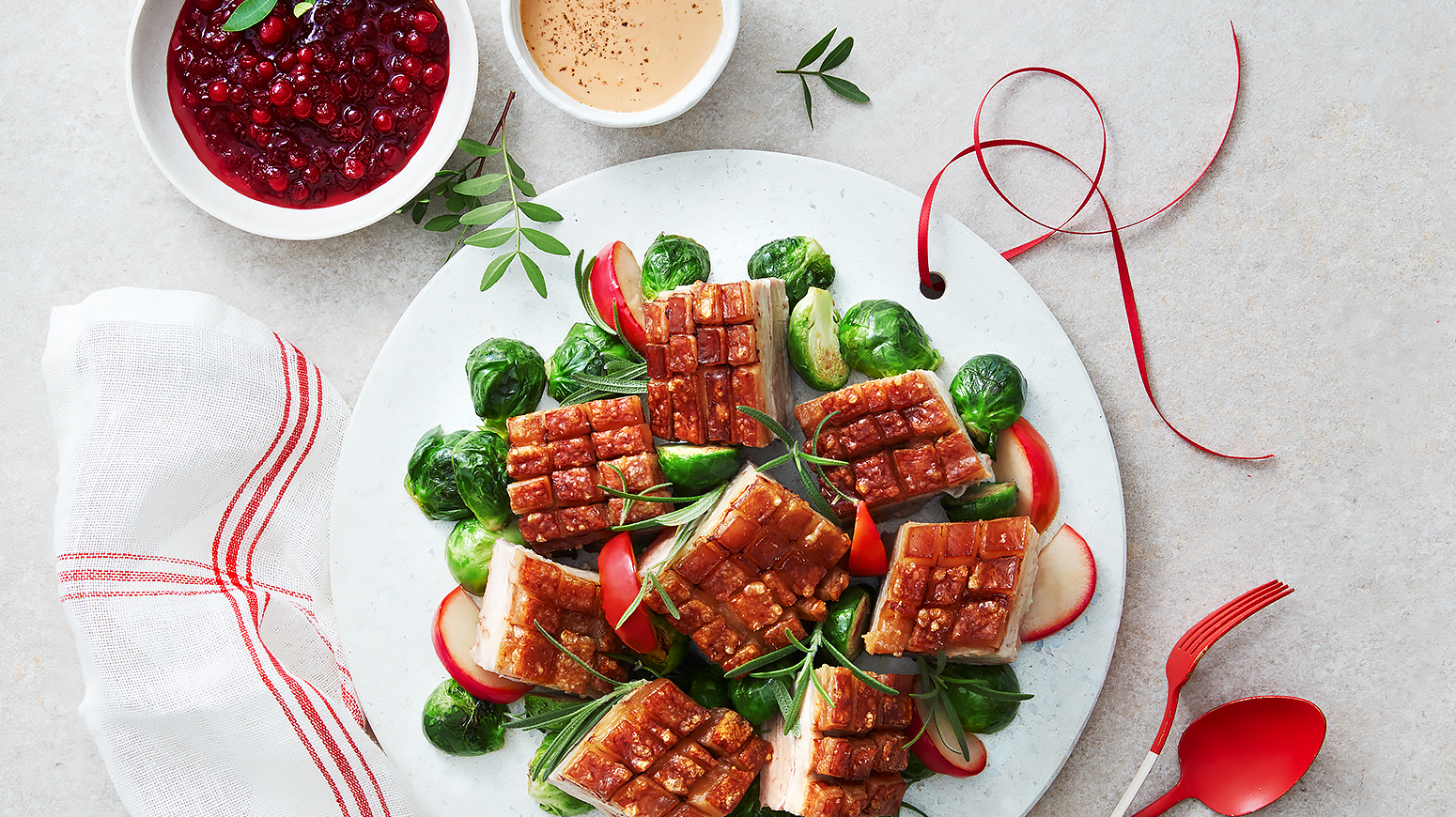 Ribbe med lun rosenkålsalat med epler, servert på hvit tallerken med rødstripet kjøkkenhåndkle og rødt bestikk ved siden av. To skåler med rørte tyttebær og fløtesaus på siden.