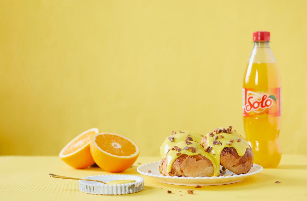 Solo-boller med Daim pyntet med gul glasur og Daimbiter, servert på hvit tallerken på gult bord med en flaske Solo i bakgrunnen. Fersk appelsin delt i to ved siden av.