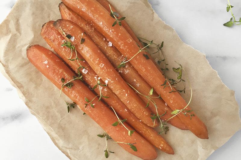 Hele honningbakte gulrøtter med flaksalt og fersk timian ligger på naturfarget bakepapir på hvit marmorbakgrunn.