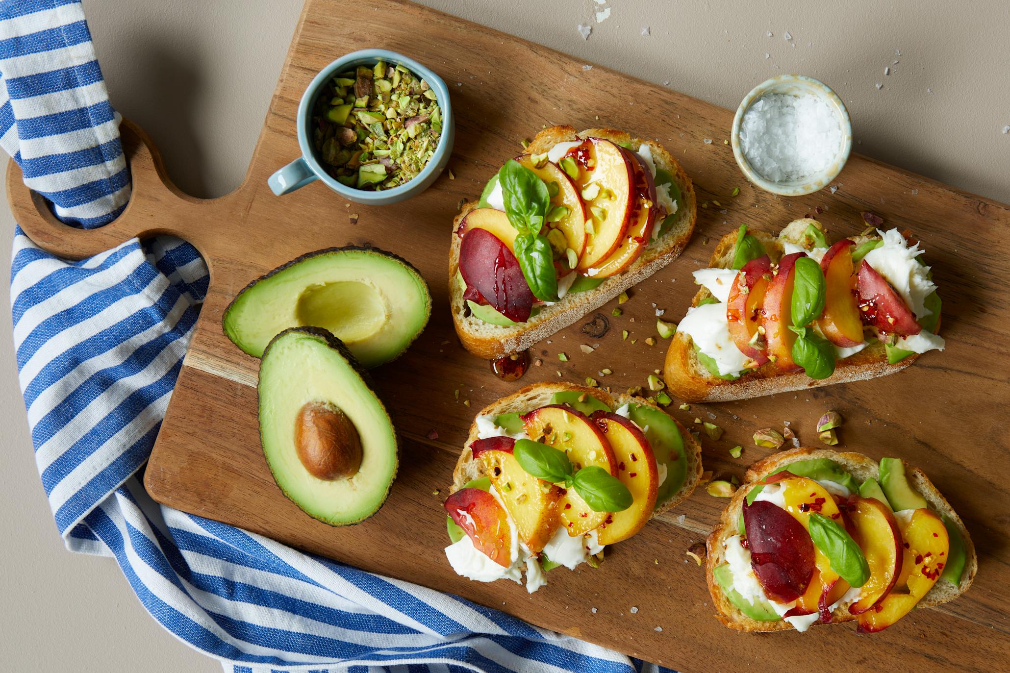 Avokadotoast med mozzarella, grillet fersken og hot honey servert på firkantet trefjøl med frisk basilikum og et dryss pistasjenøtter. To halve avokado og to små skåler med pistasjenøtter og flaksalt ved siden av.