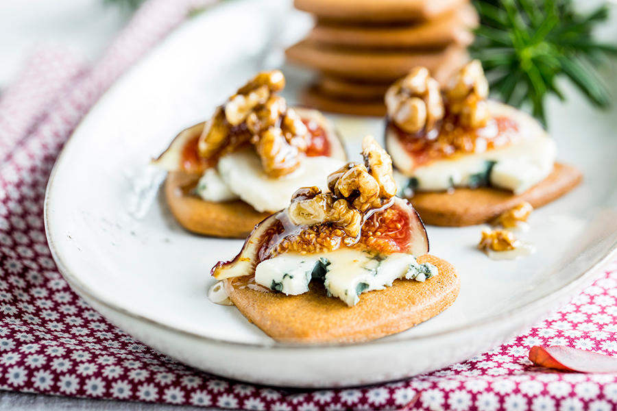 Pepperkaker med blåmuggost, fiken og honningnøtter servert på et hvitt fat på rød juleduk, med granbar i bakgrunnen.
