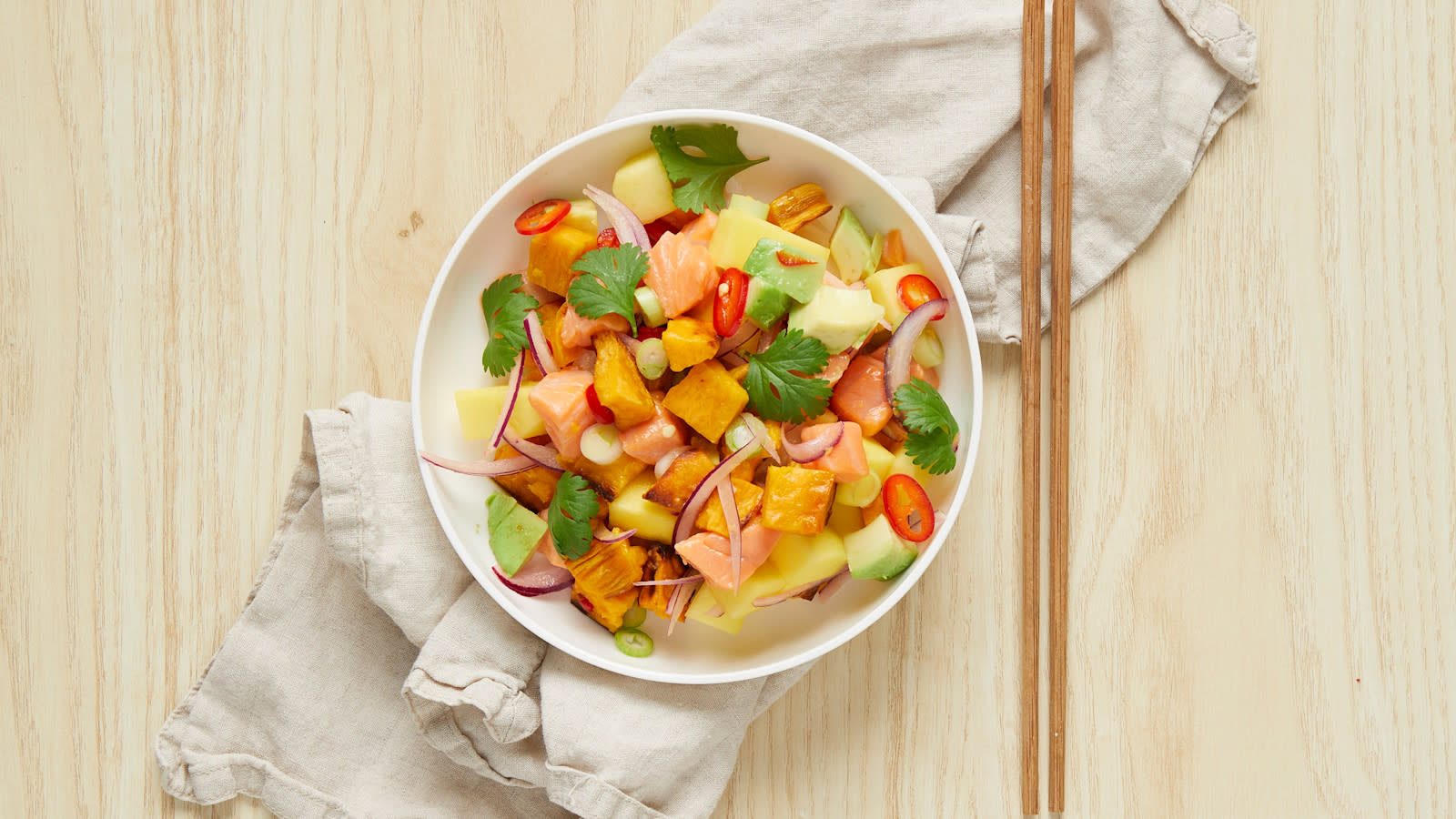 Ceviche av laks med mangosalsa og ovnsbakt søtpotet, servert i hvit skål på linserviett. Frisk koriander på toppen og spisepinner i bambus ved siden av.