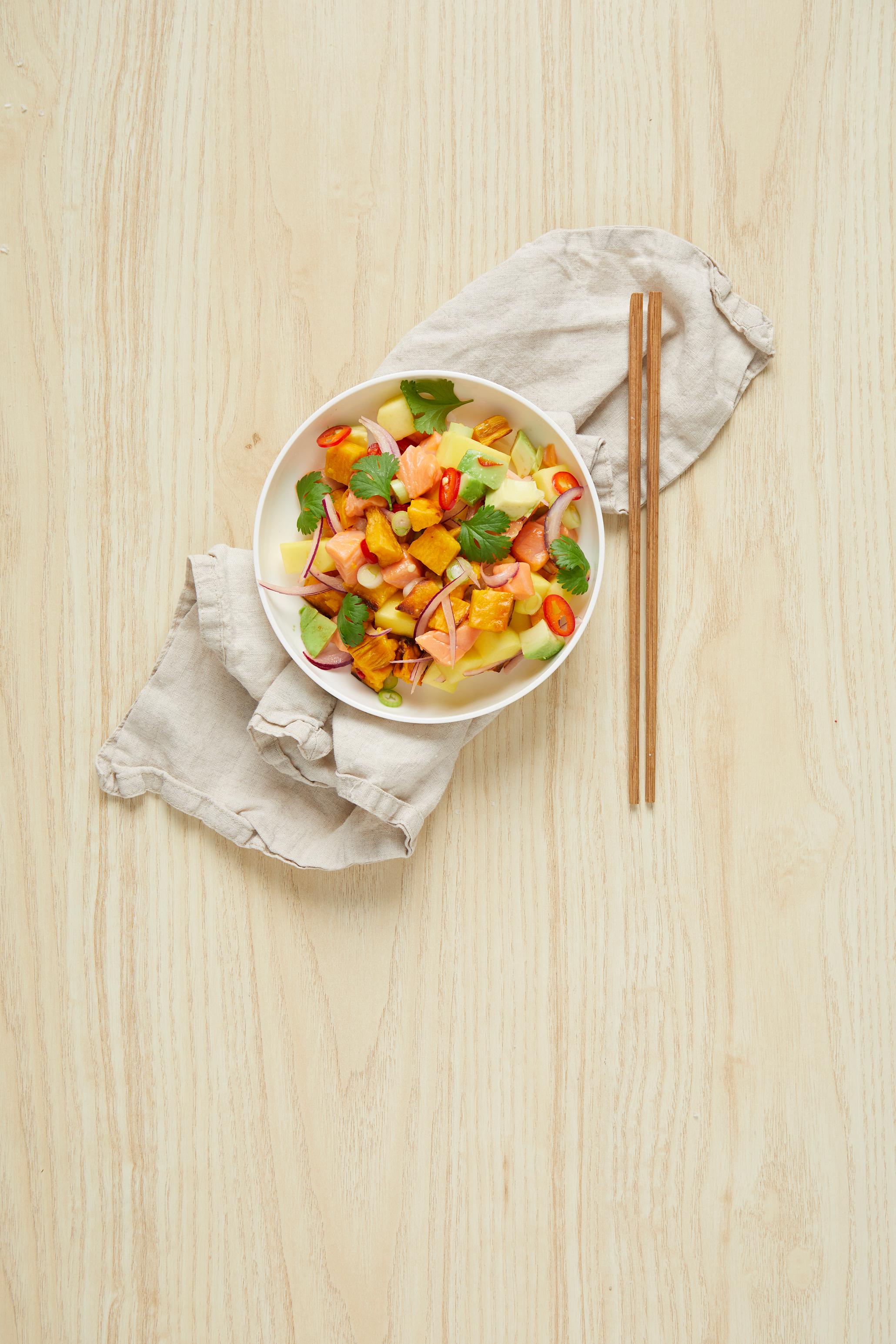 Ceviche av laks med mangosalsa og ovnsbakt søtpotet, servert i hvit skål på linserviett. Frisk koriander på toppen og spisepinner i bambus ved siden av.