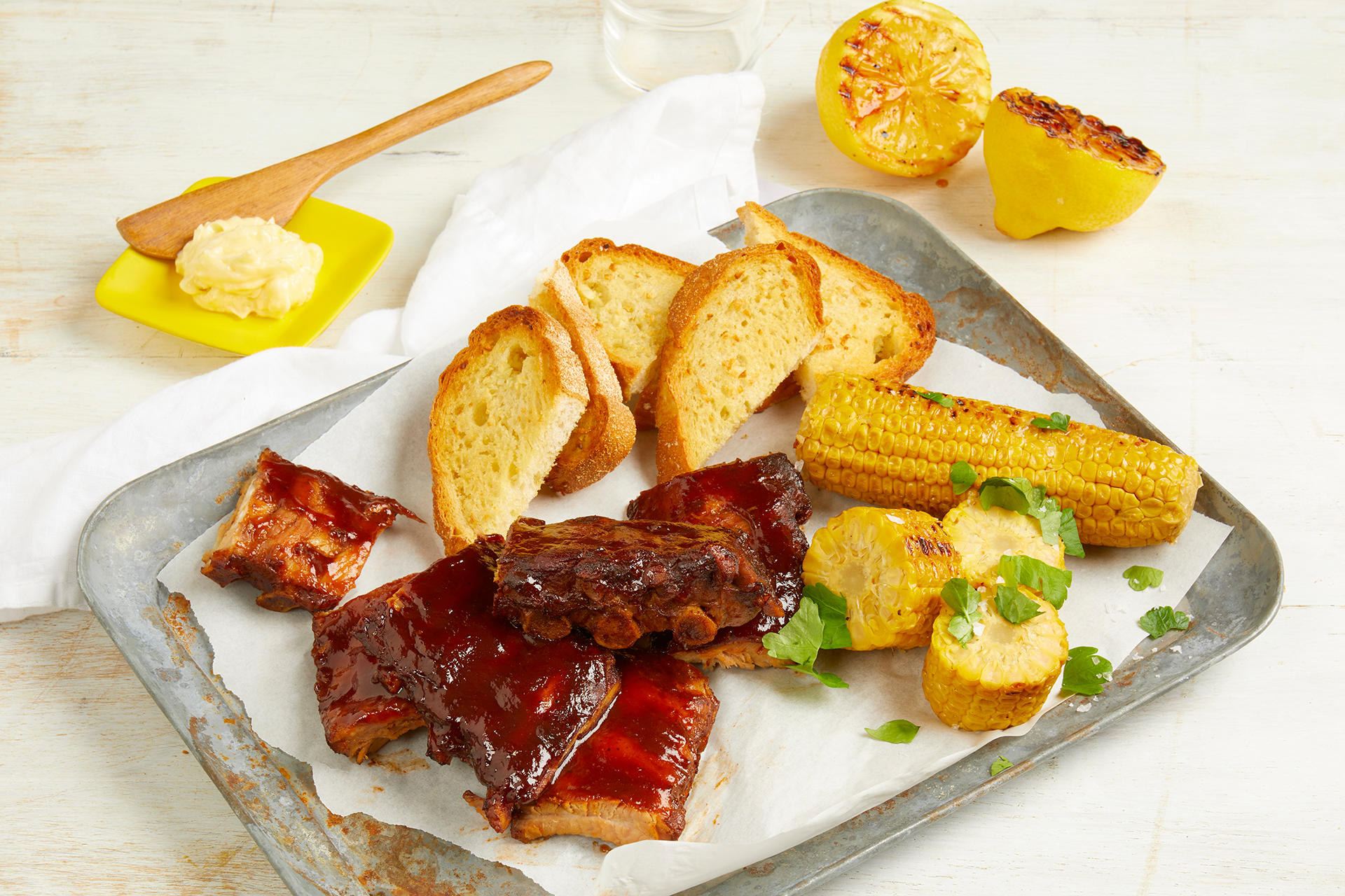 Sticky & Smokey Spareribs med hvitløkstoast og grillet mais, servert på metallbrett med hvitt bakepapir. Liten gul skål med hjemmelaget hvitløkssmør i bakgrunnen, og grillede halve sitroner på siden.
